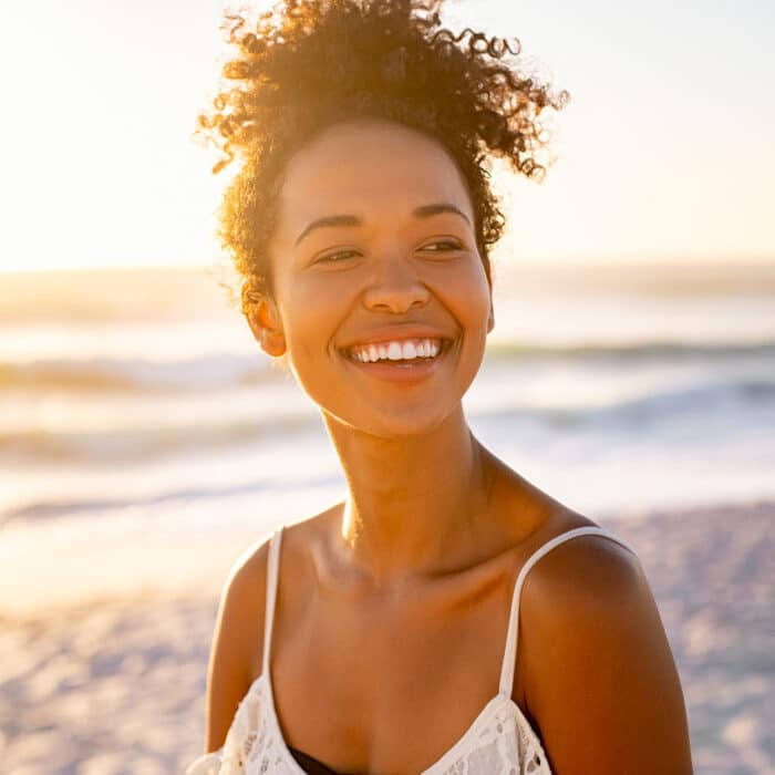 Smiling young black woman in beachwear enjoy sunset at beach. Satisfied beautiful girl with afro hair relaxing at beach during sunrise with copy space. African american woman daydreaming during sunrise.