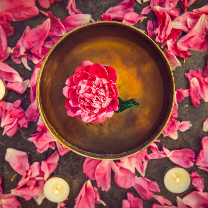 Top view tibetan singing bowl with floating inside in water pink peony flower. Burning candles and petals on the black stone background. Meditation and Relax. Exotic massage. Flatlay. Selective focus.