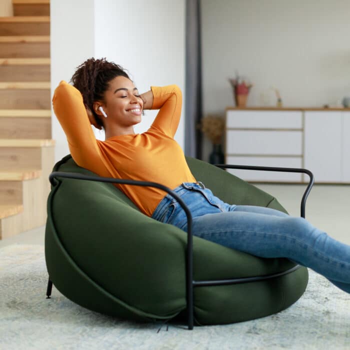 Rest And Relax Concept. Calm black woman sitting on bean bag, listening to music, audio book, podcast, enjoying meditation for sleep and peaceful mind in wireless earphones, leaning back, copy space
