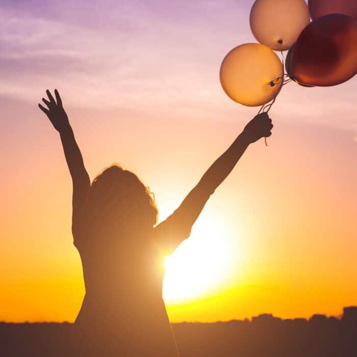 Silhouette of joyful woman with hands up holding bunch of bright balloons while standing against sunset sky