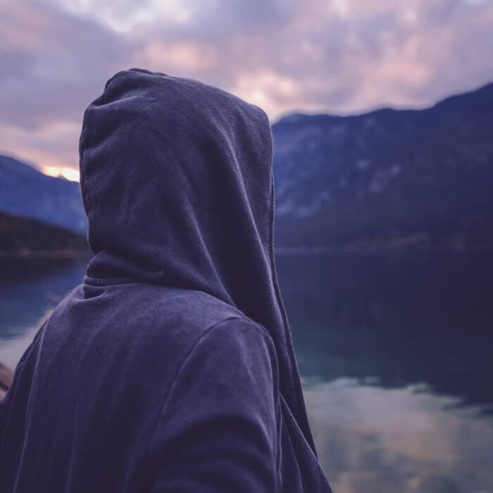 Lonely hooded female person standing by the lake and looking at beautiful mountain and forest landscape in sunset, ultra violet toned shadows