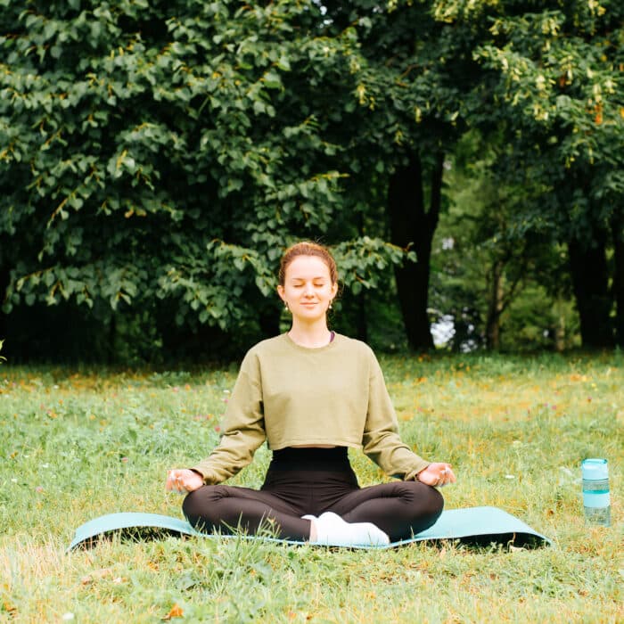 Meditation, mindfulness and spirituality concept. Young woman in sportswear meditating in lotus position with closed eyes, practicing yoga asana on sports mat in park outdoors. Sports and recreation.