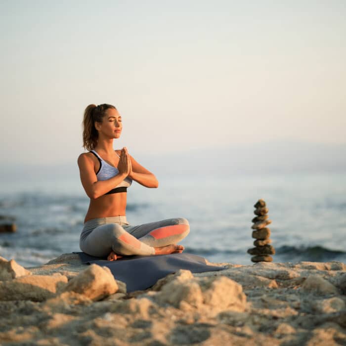 Young woman with hands clasped meditating on beach rock during morning Yoga class. Copy space.