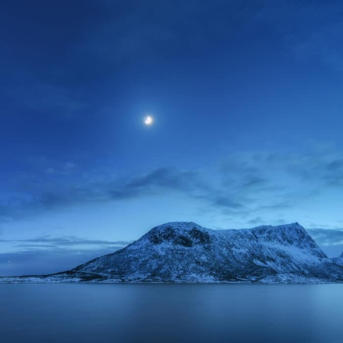 Snow covered mountains against blue sky with clouds and moon in winter at night in Lofoten islands, Norway. Arctic landscape with sea, snowy rocks, moonlight, reflection in water. Beautiful fjord