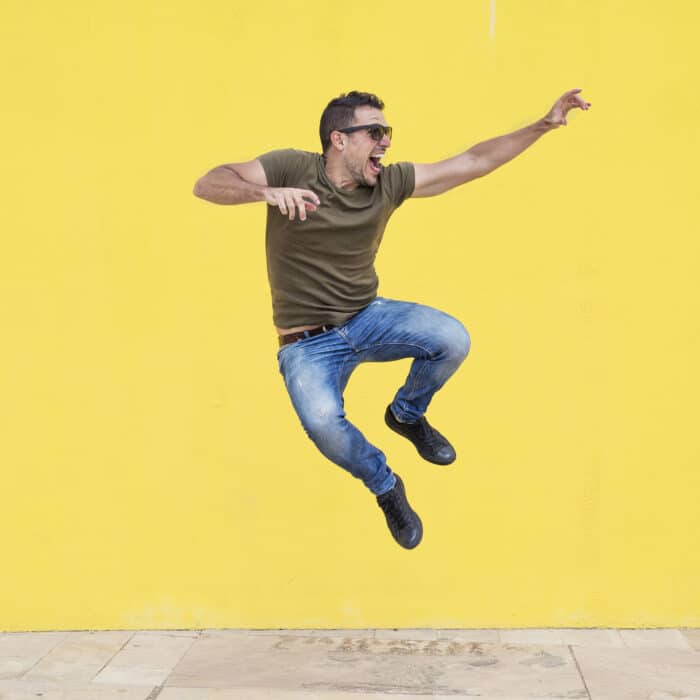 Young man with sunglasses jumping in front of a yellow wall.