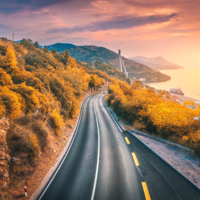Aerial view of mountain road and beautiful orange forest at colorful sunset in autumn. Dubrovnik, Croatia. Top view of road, trees, sea, mountain, red sky in fall. Landscape with highway and sea coast