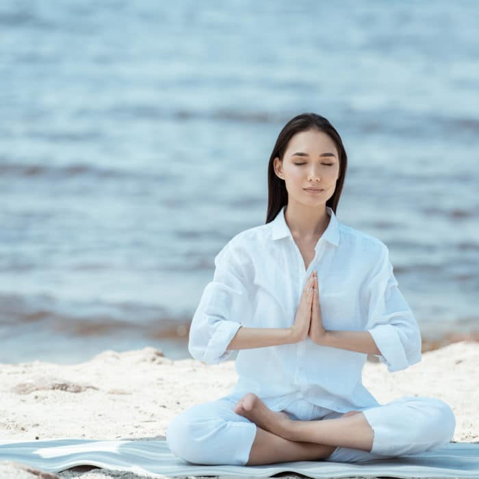 Attractive asian woman meditating in anjali mudra (salutation seal) pose on yoga mat by sea