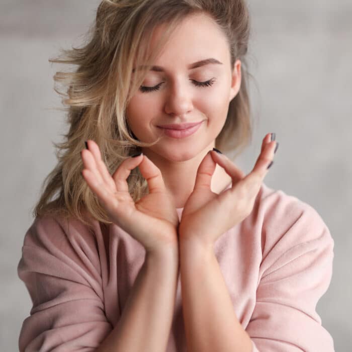 Charming young blond woman in pink sweatshirt holding eyes closed and holding hands in meditation pose in daylight