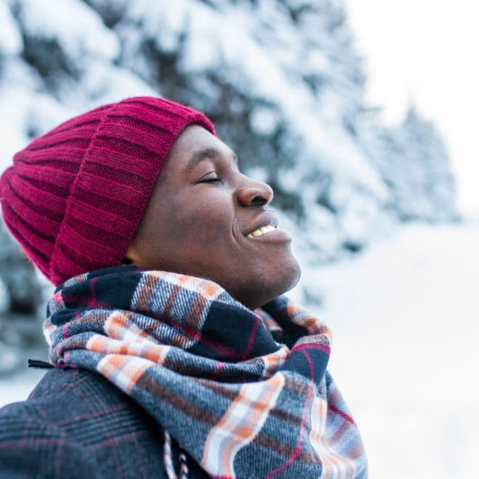 Happy carefree african american man in warm clothes taking a deep breath in fir forest .
