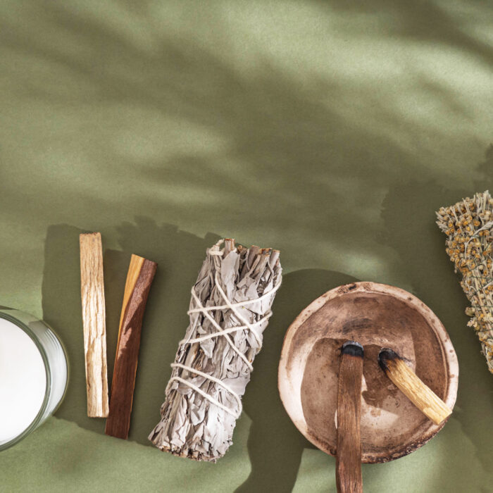 Items for spiritual cleansing - sage and various herbs bundles, palo santo incense sticks and candle on green background with shadows. Top view. Copy space