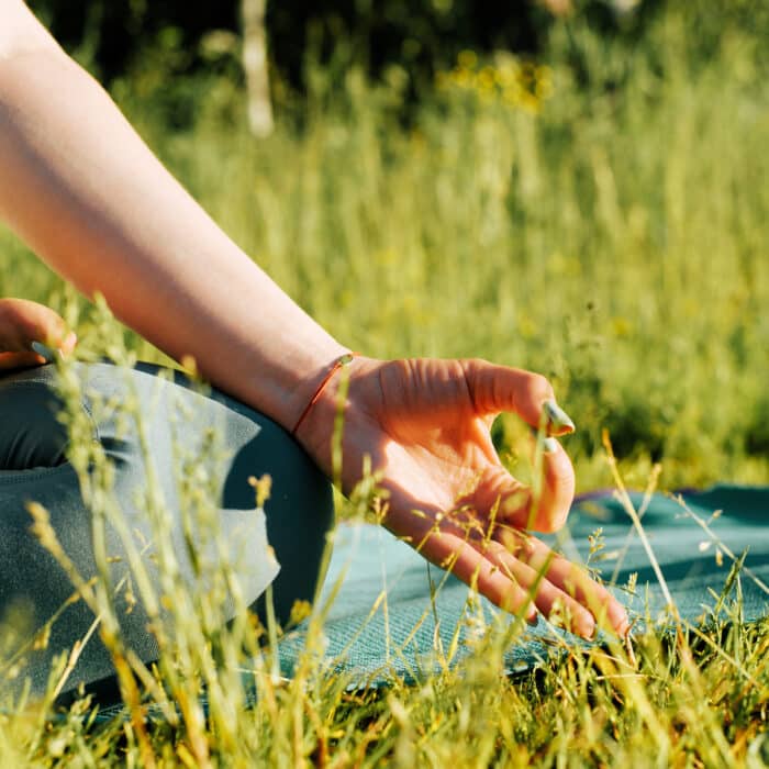 Meditation, outdoors. Close-up of a female hand in a yoga asana pose for Gyan Mudra meditation. Woman meditating in lotus position sitting on a rug on green grass in nature, cropped image.