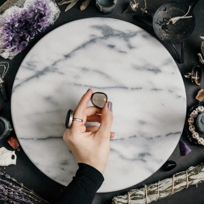 Witch's hand holding Smoky quartz above a marble white round tray. The place for witchcraft with magic things around. View from above.