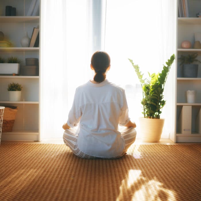 Young woman enjoying sunny morning and practicing meditation near window at home.