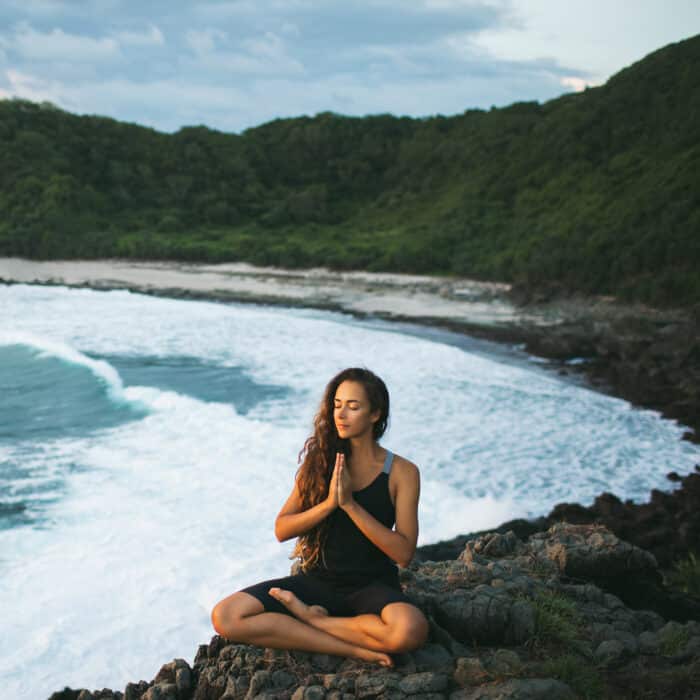 Young woman praying and meditating alone at sunset with beautiful ocean and mountain view. Self-analysis and soul-searching. Spiritual and emotional concept. Introspection, introversion and soul healing.