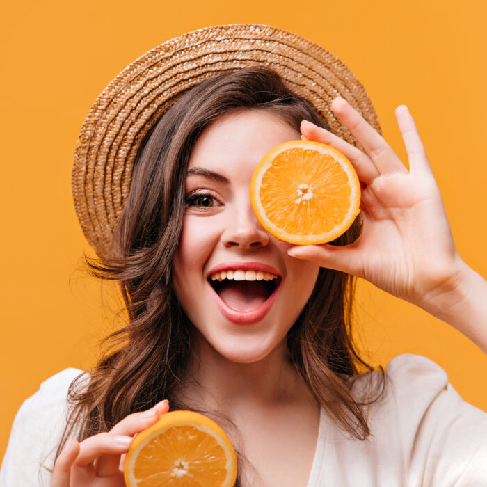 Optimistic woman in straw hat covers her eyes with orange and smiles while looking at camera
