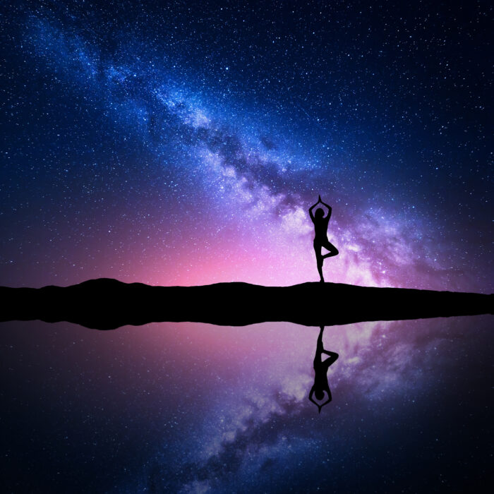 Milky Way. Silhouette of a standing woman practicing yoga on the mountain near the pond with sky reflection in water. Landscape with meditating girl on the hill. Night starry sky and milky way