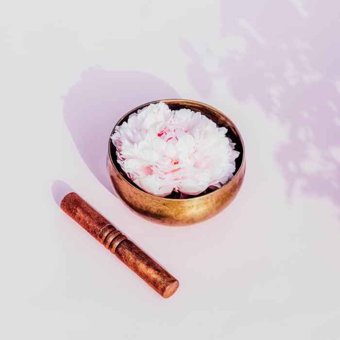 Tibetan singing bowl with floating in water pink peony inside and wooden stick on the pink background. Meditation and Relax. Exotic massage. Minimalism. Direct sunlight and shadows. Selective focus