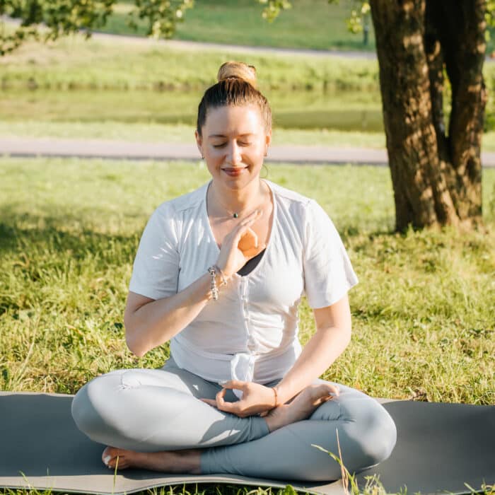 Yoga and meditation, mind health and mental harmony concept. Calm smiling young yogi woman sitting in lotus position on sports mat, practicing meditation in park at sunrise. Active healthy lifestyle