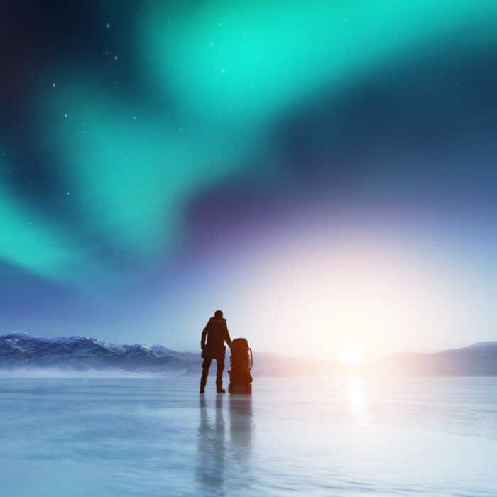 Adventurous man standing on a frozen lake with a backpack, looking at northern lights. Travel and adventure.