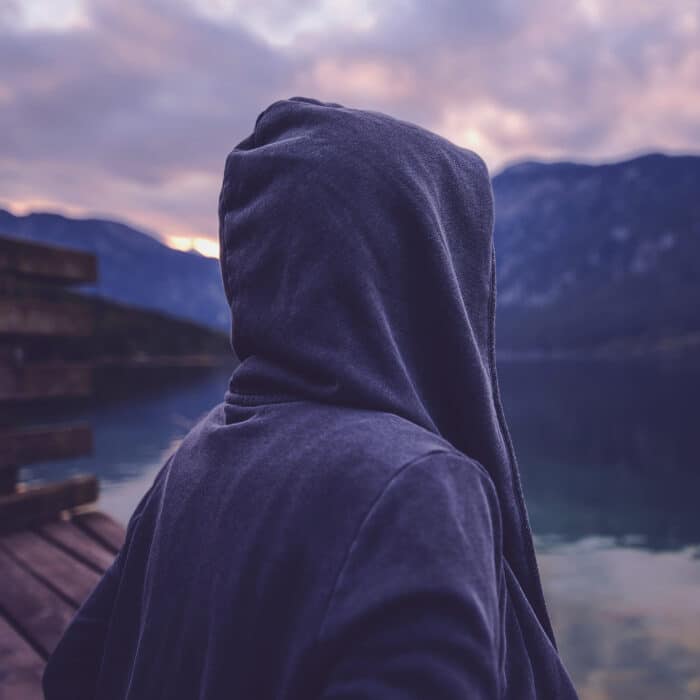 Lonely hooded female person standing by the lake and looking at beautiful mountain and forest landscape in sunset, ultra violet toned shadows