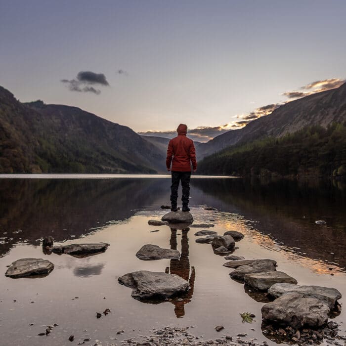 A male in an orange jacket standing on a stone in the middle of Glendalough Lake in Wicklow, Ireland