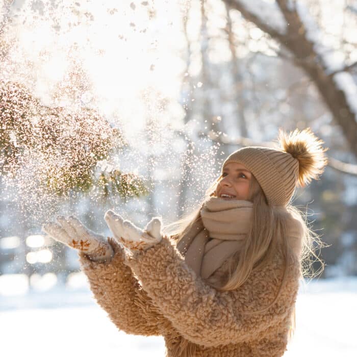 Attractive caucasian blonde woman with long hair throws snow up with a nice smile, bottom view. Happy young female plays with a snow in sunny winter day. Winter time concept