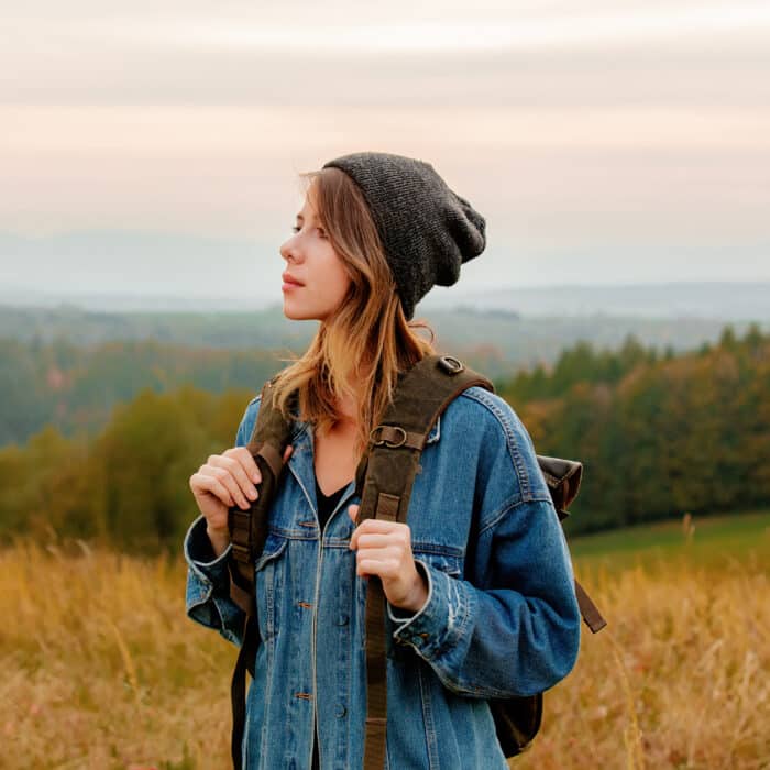 Style girl in denim jacket and hat with backpack in countryside with mountains on background