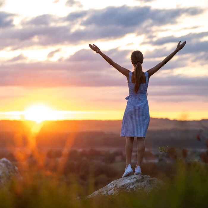 Dark silhouette of a young woman standing with raised up hands on a stone enjoying sunset view outdoors in summer.