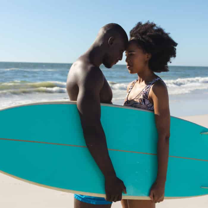 Happy african american couple holding surfboard standing on sunny beach touching heads. Summer, togetherness, romance, healthy lifestyle, sport, hobbies, surfing and vacation, unaltered.