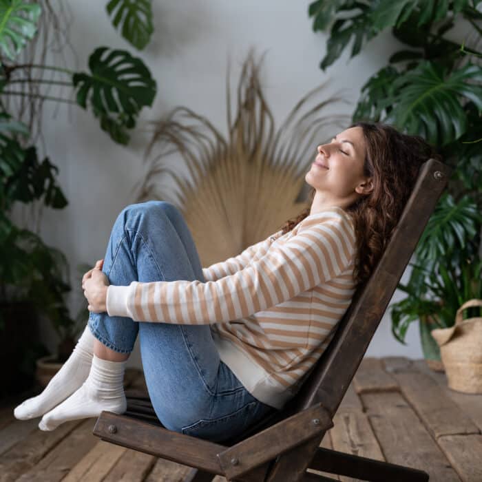 Happy calm peaceful Polish woman sitting on wooden chair dreaming with closed eyes, resting in urban jungle interior, dreamy serene Hispanic female relaxing in tropical greenhouse. Daydreaming