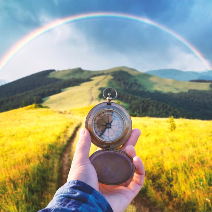 Man with old metal compass in hand on mountains road. Travel concept. Lush green grassy meadows and beautyful rainbow on background. Landscape photography