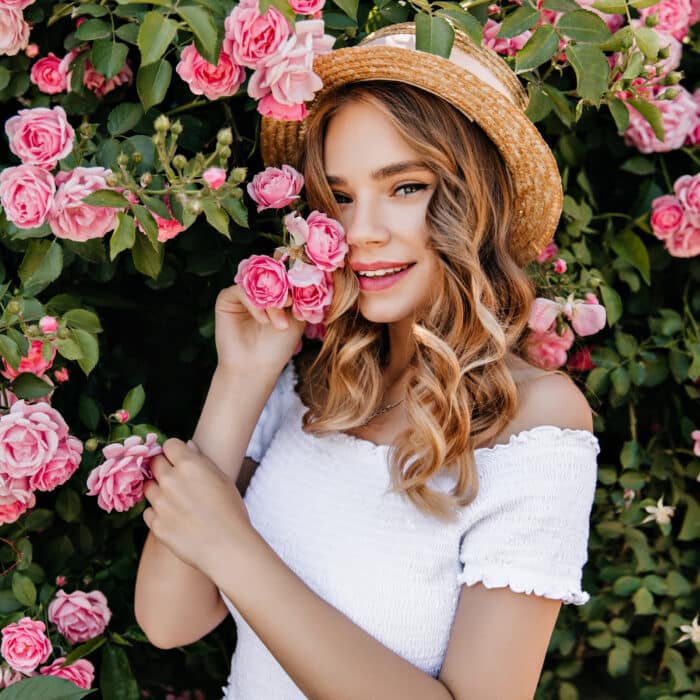 Outdoor portrait of happy white girl posing on nature background. Photo of relaxed lady with wavy hair standing near beautiful rose bush.