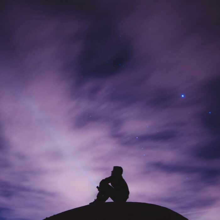 A beautiful wide shot of a person sitting on a rock under a purplish starry night sky