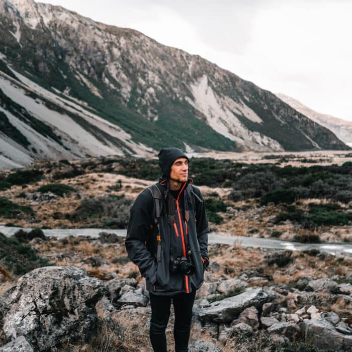 Happy and calm young caucasian man enjoying the landscape with a black hoodie a camera hanging and hands in pockets on a very cold day under a gray sky near the snowy mountains and on the rocks, mount cook, new zealand - Travel concept