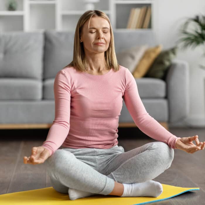 Meditation Concept. Beautiful Calm Woman Meditating At Home In Lotus Position, Peaceful Young Caucasian Lady Practicing Yoga, Sitting With Closed Eyes On Fitness Mat In Light Room, Copy Space