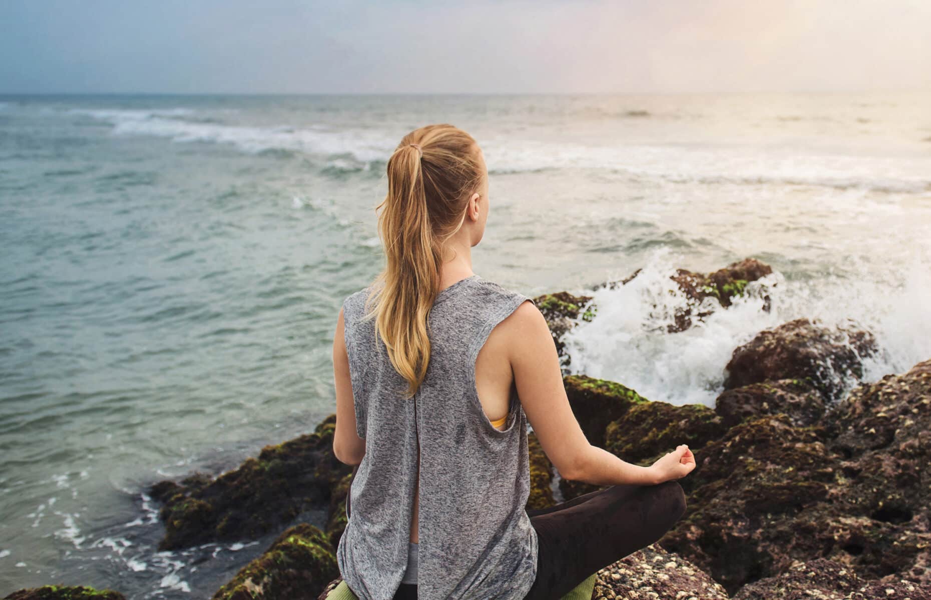 Woman meditate on the rock near the sea with wave splashes on sunset