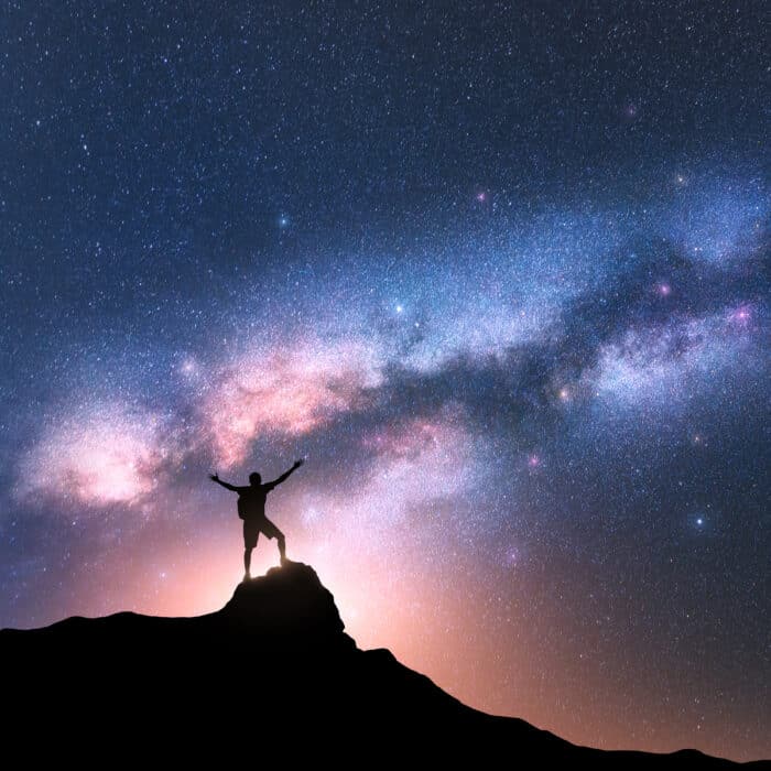 Milky Way and happy man with backpack on the mountain peak at night. Silhouette of guy with raised up arm on the hill, sky with stars, yellow light in Nepal. Galaxy. Space landscape with milky way