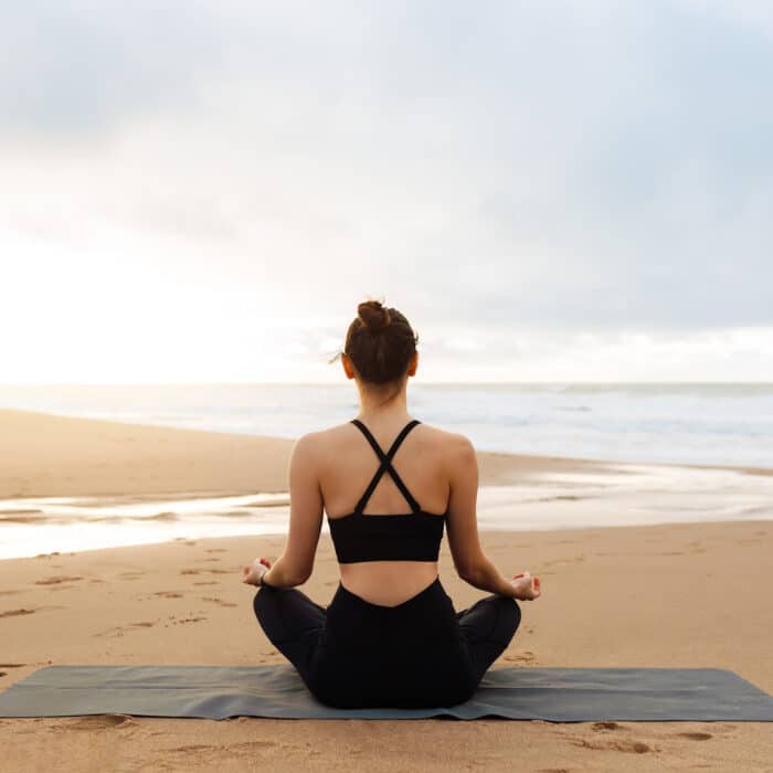 Morning meditation. Unrecognizable calm woman in activewear practicing yoga outdoors, lady sitting in lotus position on fitness mat on beach near ocean, enjoying meditating