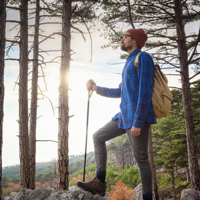 The concept of discovery and hiking, nature and freedom. Young man standing on top of a cliff in summer mountains among pine forest and enjoying nature view