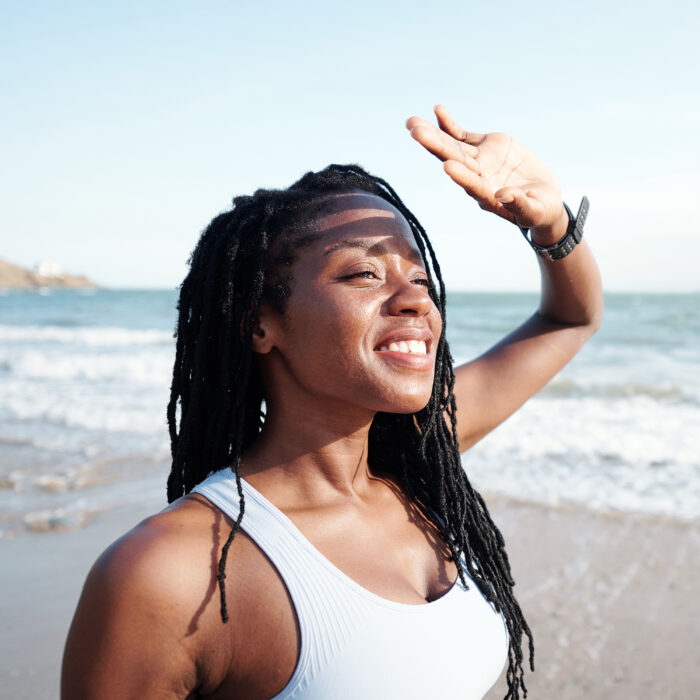 Cheerful young woman covering her eyes with hands from bright sunlight when looking at seascape