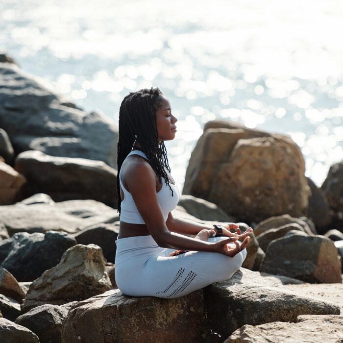 Calm relaxed sportswoman sitting on rocky beach, enjoying sea breeze and meditating